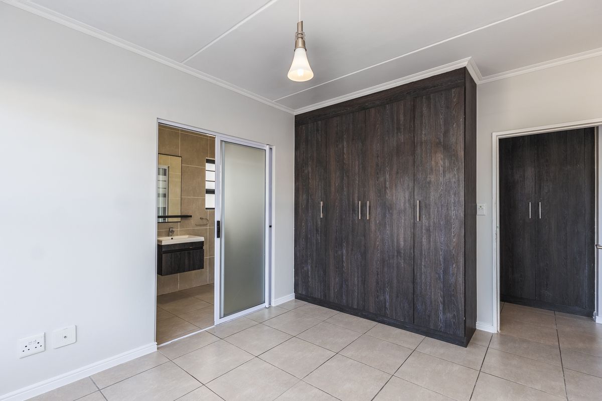 A clean, modern bedroom featuring dark wood built-in cupboards, neutral tiled flooring, and an en-suite bathroom accessible via a sliding door.