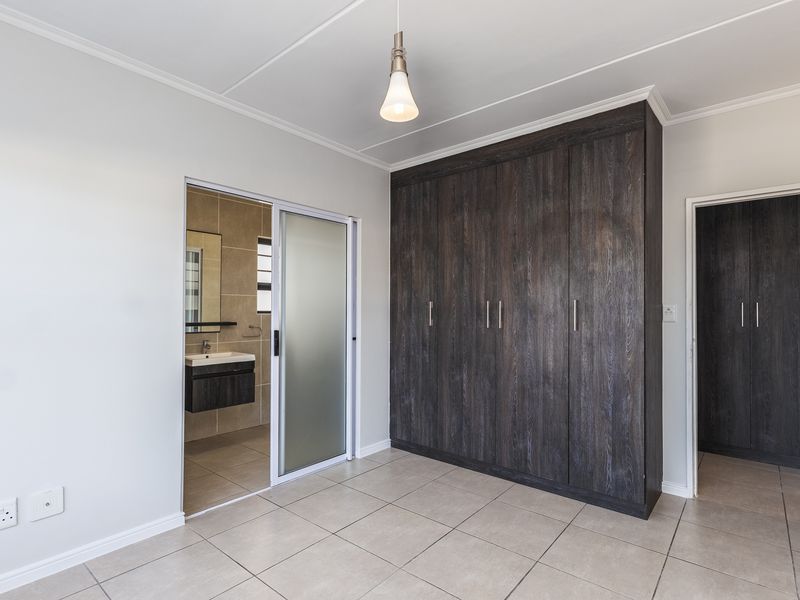 A clean, modern bedroom featuring dark wood built-in cupboards, neutral tiled flooring, and an en-suite bathroom accessible via a sliding door.