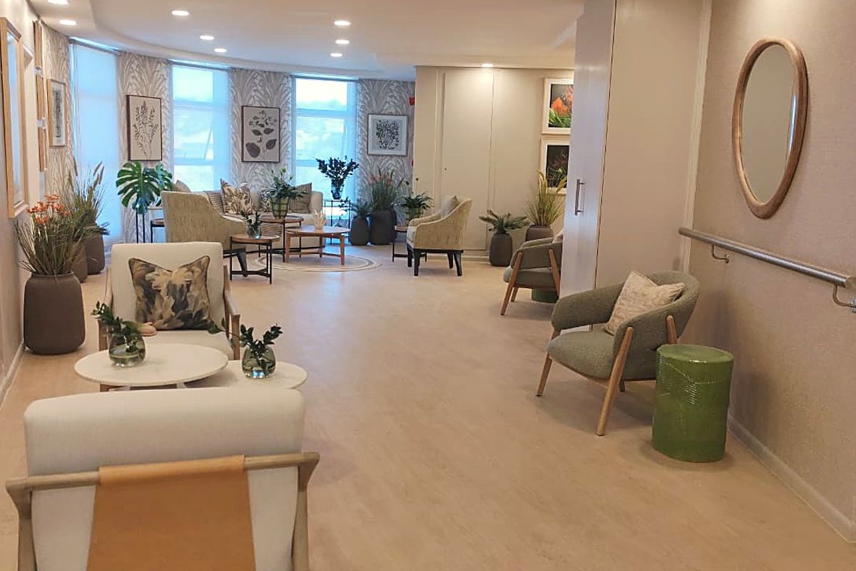 A spacious, well-lit communal living area featuring neutral vinyl flooring, recessed lighting, and a wall-mounted grab rail for accessibility.