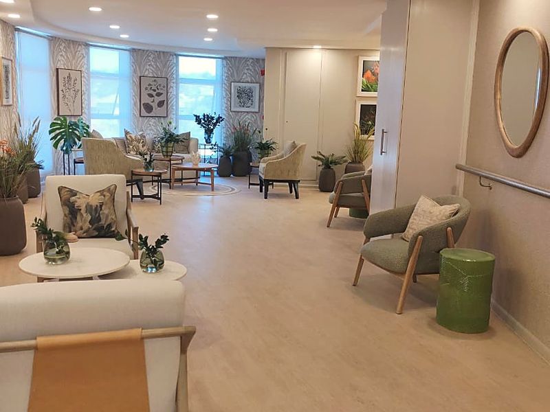 A spacious, well-lit communal living area featuring neutral vinyl flooring, recessed lighting, and a wall-mounted grab rail for accessibility.
