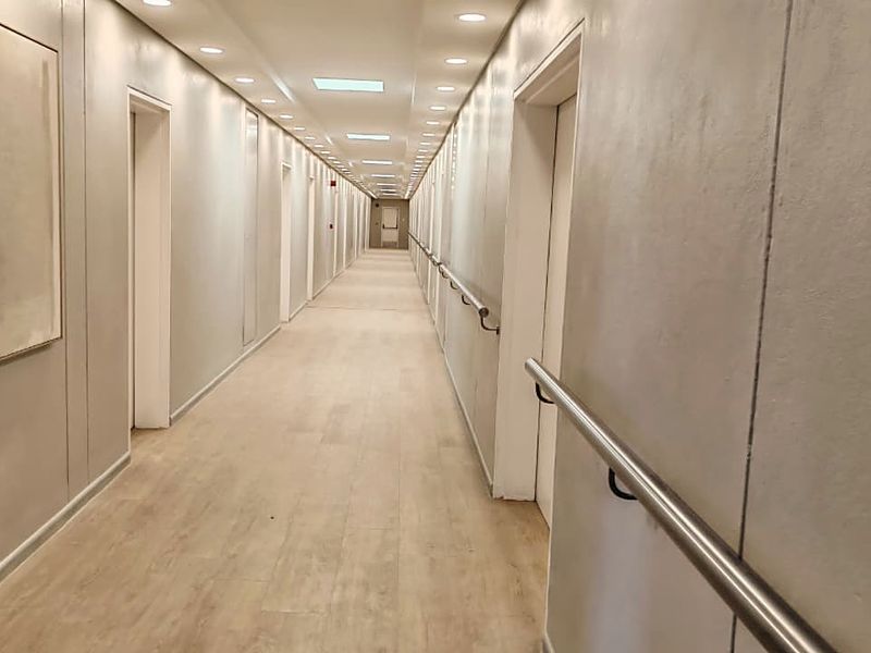 A long, well-lit apartment building hallway featuring neutral wall finishes, laminate flooring, and safety handrails along the corridor.