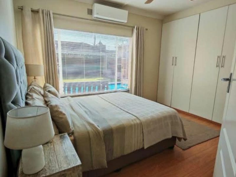 A well-maintained bedroom featuring laminate flooring, built-in cupboards, a ceiling fan, and an air conditioning unit, with a view overlooking the pool area.