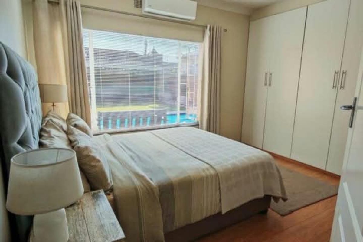 A well-maintained bedroom featuring laminate flooring, built-in cupboards, a ceiling fan, and an air conditioning unit, with a view overlooking the pool area.