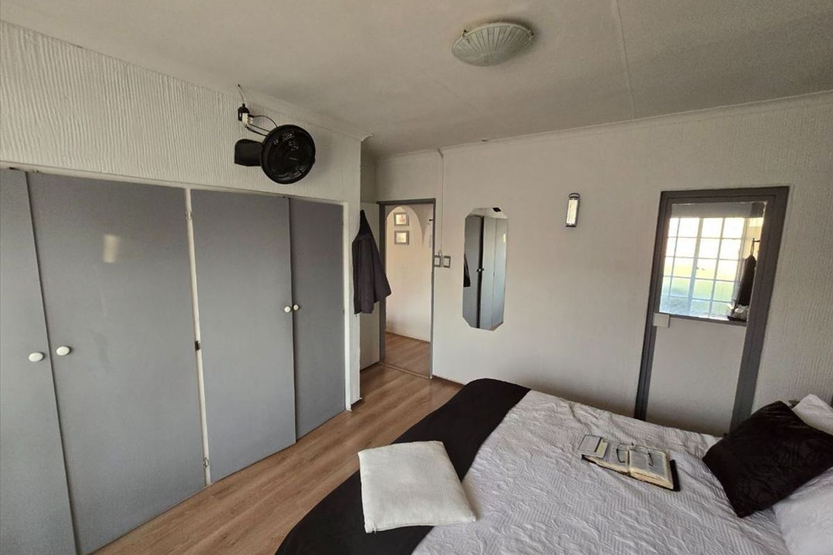 A bedroom featuring laminate flooring, built-in cupboards, and a wall-mounted fan, offering a functional space with potential for cosmetic updates.
