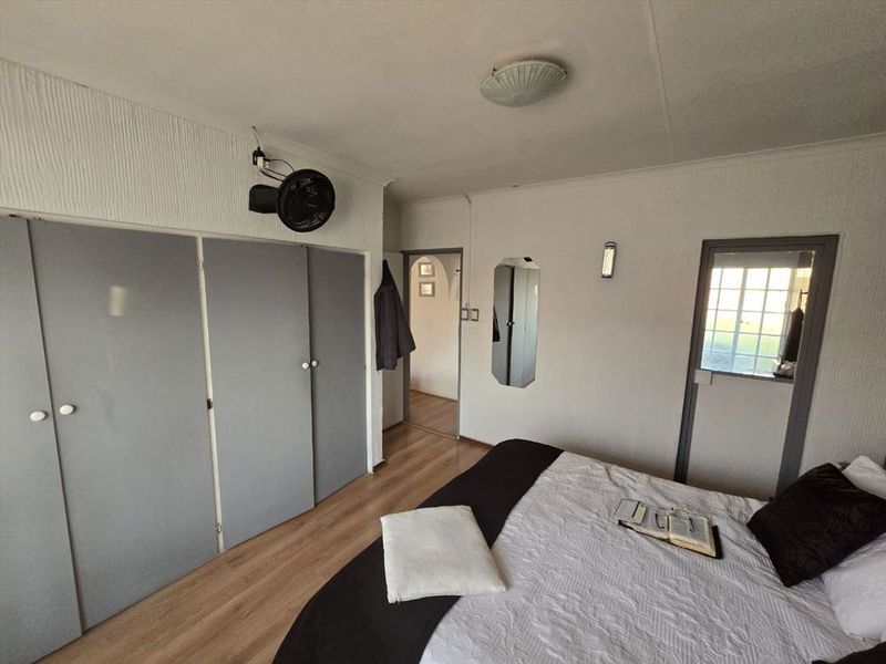 A bedroom featuring laminate flooring, built-in cupboards, and a wall-mounted fan, offering a functional space with potential for cosmetic updates.