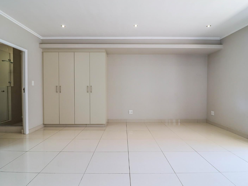 A bright, empty bedroom featuring neutral tiled flooring, built-in cupboards, and recessed downlighting, with an adjacent en-suite bathroom visible through the doorway.