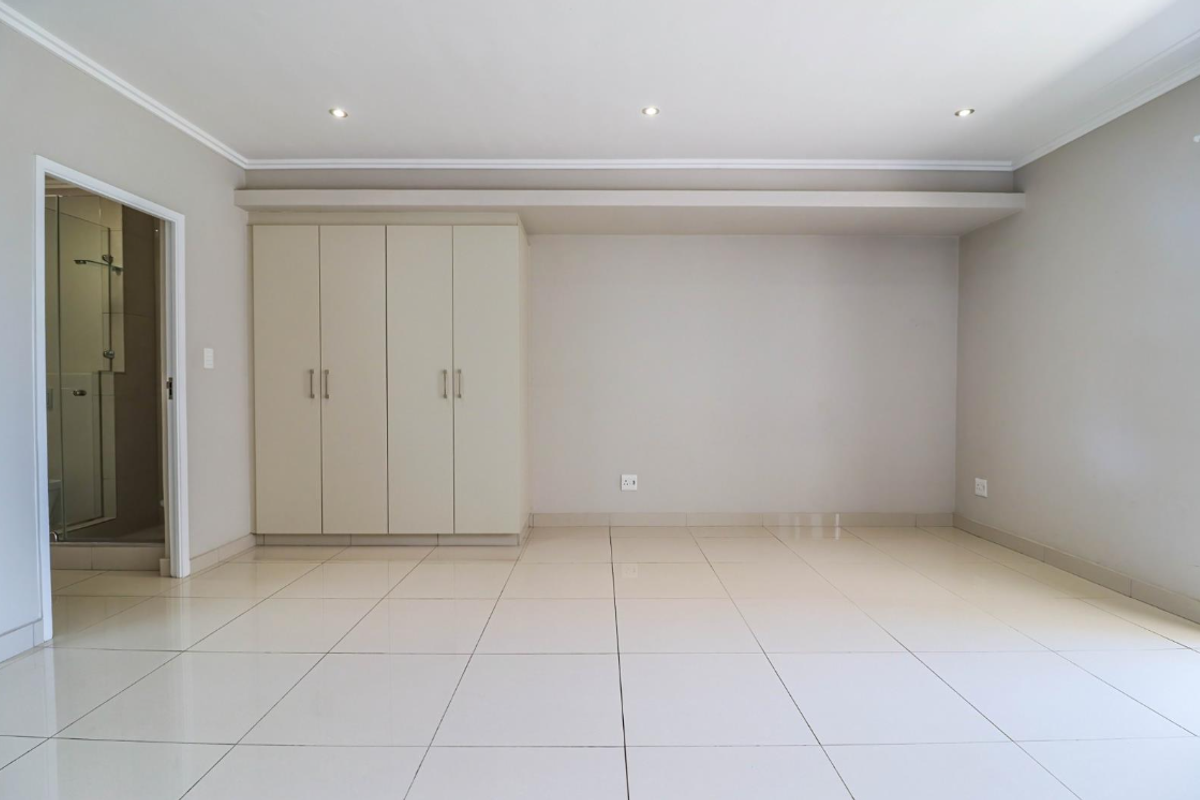 A bright, empty bedroom featuring neutral tiled flooring, built-in cupboards, and recessed downlighting, with an adjacent en-suite bathroom visible through the doorway.