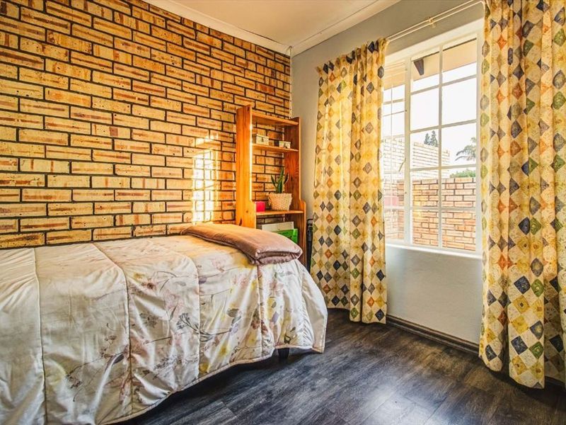 A cozy bedroom featuring a rustic exposed brick wall, durable laminate flooring, and a window with security bars allowing for natural light.