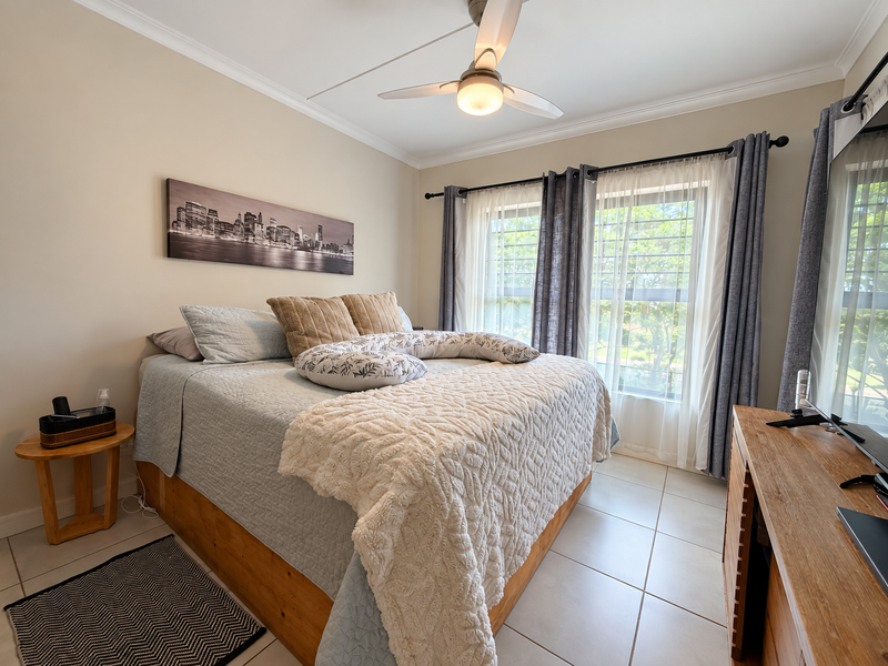 A well-maintained bedroom featuring tiled flooring, a ceiling fan, and large windows with security bars, offering a bright and comfortable living space.