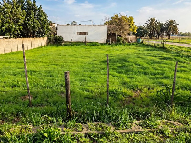 A vacant residential plot featuring a grass lawn, enclosed by wire fencing and a concrete boundary wall, with a view of the surrounding area.
