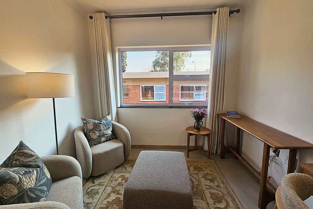A cozy living area featuring neutral tiled flooring and a large window with curtain fixtures, providing natural light and a view of the complex.
