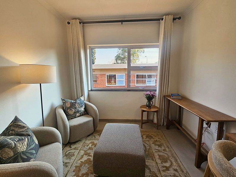 A cozy living area featuring neutral tiled flooring and a large window with curtain fixtures, providing natural light and a view of the complex.