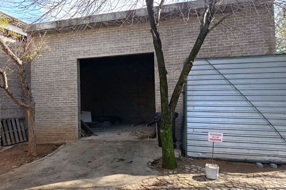 A detached brick garage structure featuring a large open bay and a corrugated metal roller door, situated on a concrete driveway.