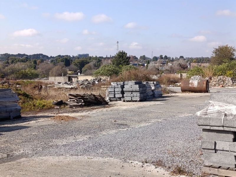 An expansive outdoor area featuring a gravel and concrete surface, currently used for storage with surrounding overgrown vegetation and distant landscape views.