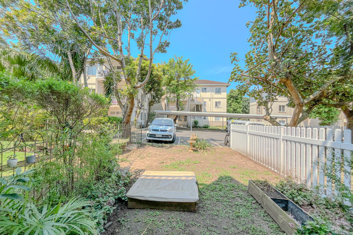 A rear garden area featuring a white picket fence, established trees, and a view towards the apartment complex parking area with covered carports.