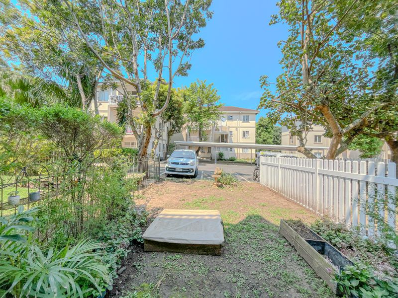 A rear garden area featuring a white picket fence, established trees, and a view towards the apartment complex parking area with covered carports.