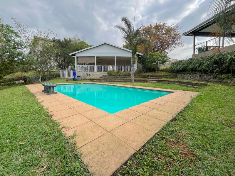 A well-maintained outdoor pool area featuring a paved surround, tiered landscaping, and a covered patio structure in the background.