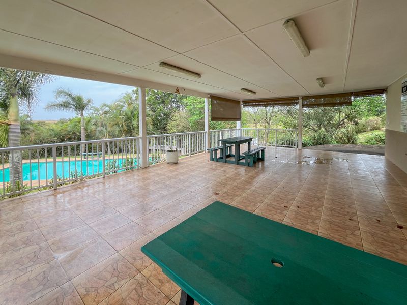 A spacious covered patio area featuring tiled flooring, protective railings, and outdoor blinds, overlooking the swimming pool and garden area.