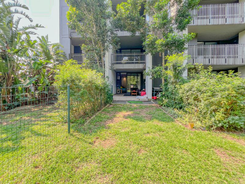 Rear view of the apartment building featuring a private garden area, tiled patio, and balcony access through sliding glass doors.