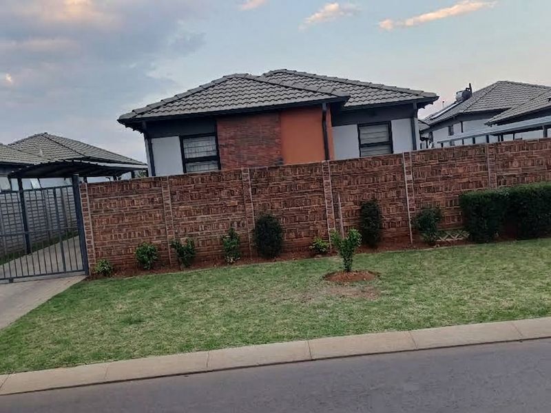 Front view of a residential property featuring a brick boundary wall, metal driveway gate, paved driveway, and a tiled roof with solar water heating.