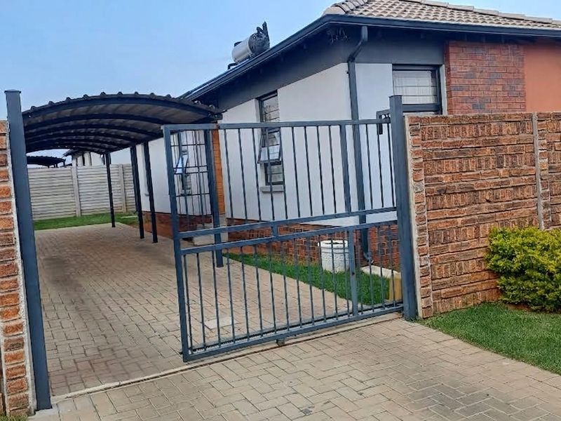Front view of the property featuring a paved driveway, covered carport, and secure metal sliding gate with brick boundary walls.