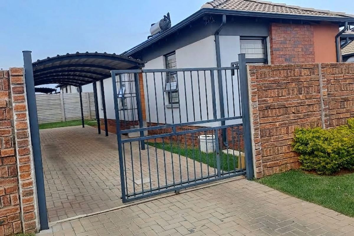 Front view of the property featuring a paved driveway, covered carport, and secure metal sliding gate with brick boundary walls.