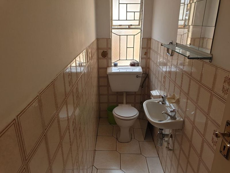 A compact bathroom featuring a standard toilet, wall-mounted basin, and full-height tiled walls with security bars on the window.