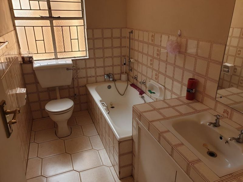 A dated bathroom featuring a built-in bathtub, toilet, and vanity sink with original tiled finishes and security bars on the window.