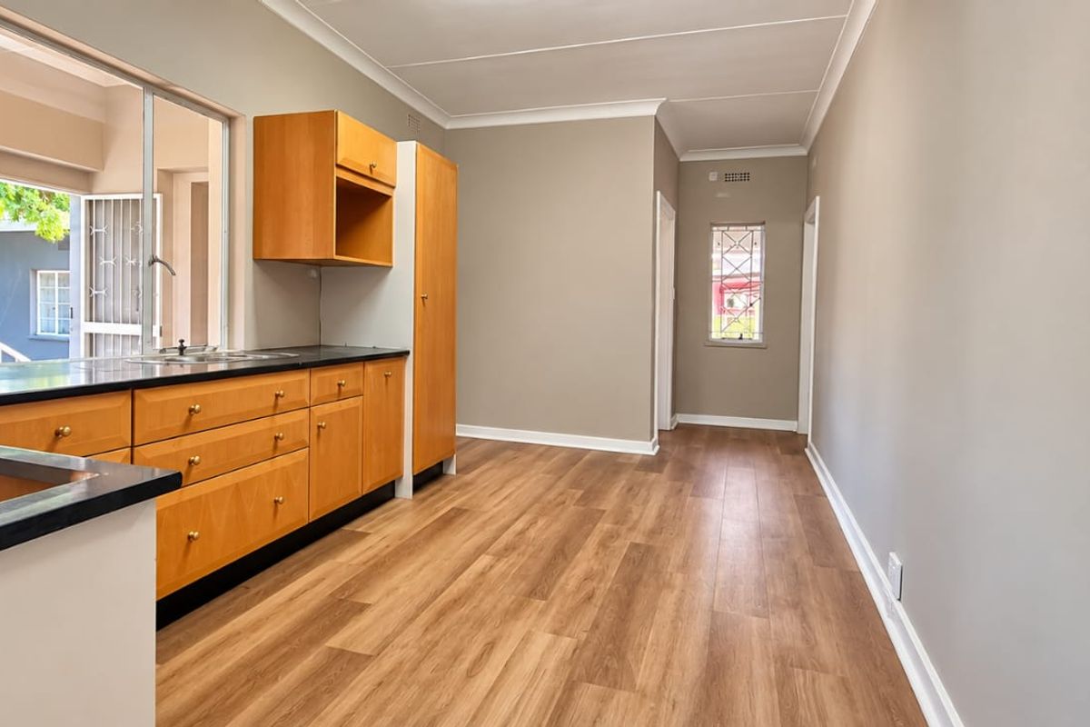 A clean, empty kitchen featuring warm-toned wooden cabinetry, dark laminate countertops, and modern wood-look flooring with ample natural light.