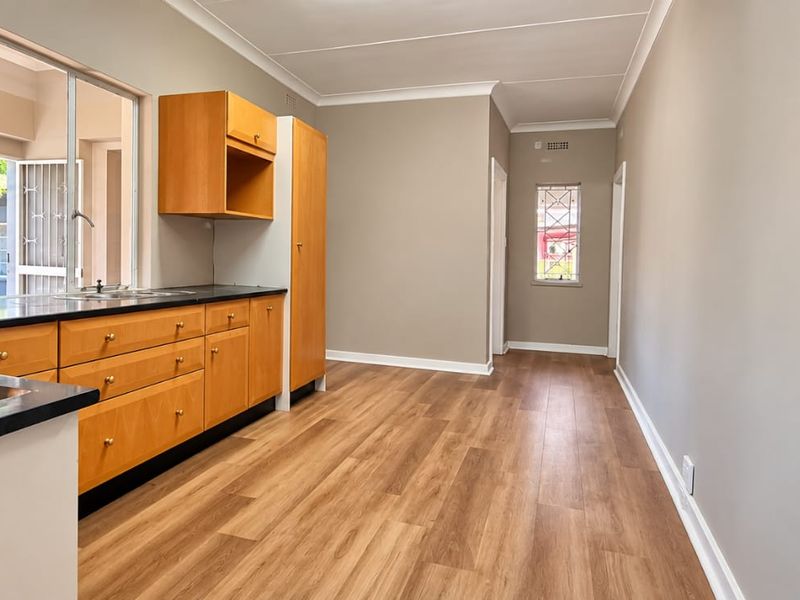 A clean, empty kitchen featuring warm-toned wooden cabinetry, dark laminate countertops, and modern wood-look flooring with ample natural light.
