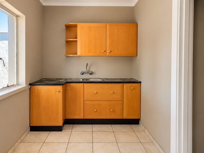 A functional laundry room featuring built-in wooden cabinetry, a stainless steel sink, and neutral tiled flooring with natural light from a side window.