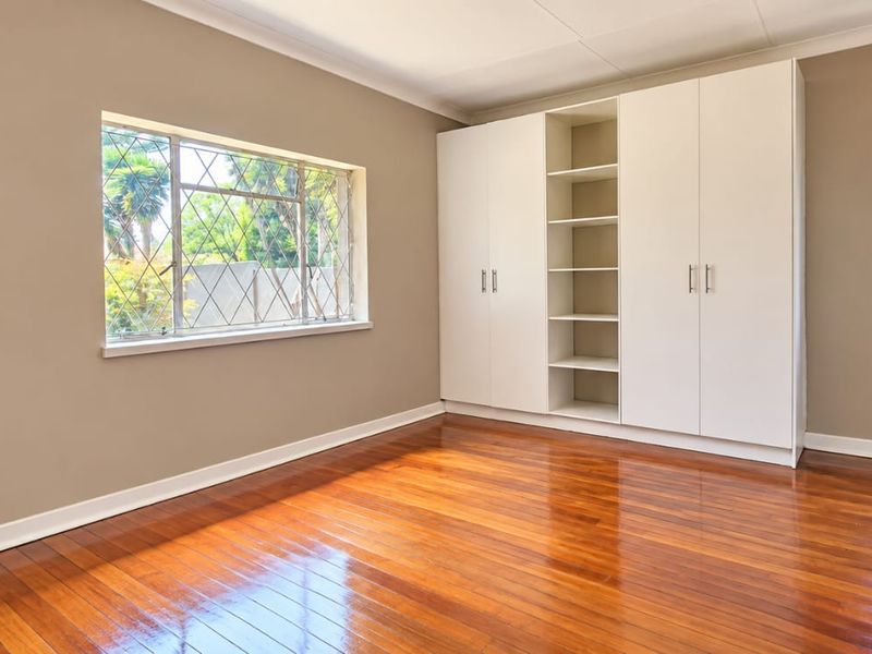 A bright, empty bedroom featuring polished hardwood flooring, neutral wall tones, and extensive built-in cupboard storage with integrated shelving.