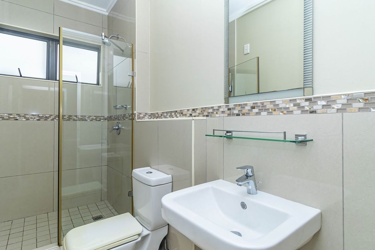A clean, modern bathroom featuring a glass-enclosed shower, wall-mounted basin with chrome fixtures, and neutral tiled walls with a decorative mosaic accent border.