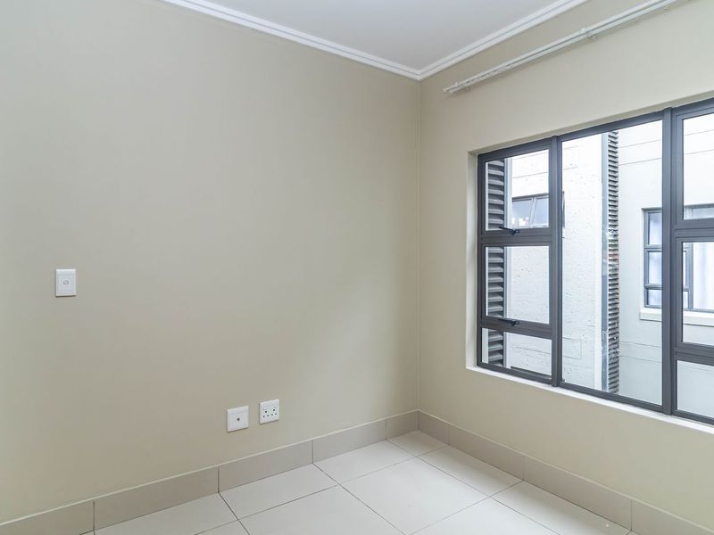 A clean, empty bedroom featuring neutral wall paint, tiled flooring, and large aluminium-framed windows allowing natural light.