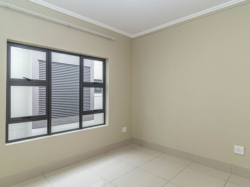 A clean, empty bedroom featuring neutral wall tones, tiled flooring, and large aluminium-framed windows providing natural light.