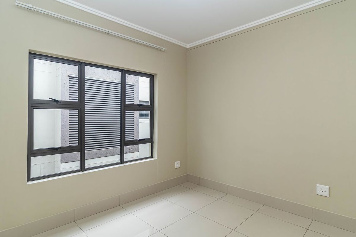 A clean, empty bedroom featuring neutral wall tones, tiled flooring, and large aluminium-framed windows providing natural light.