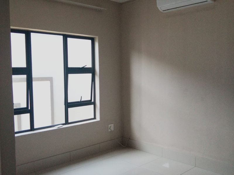 A clean, empty bedroom featuring neutral tiled flooring, an installed air conditioning unit, and large aluminium-framed windows providing natural light.