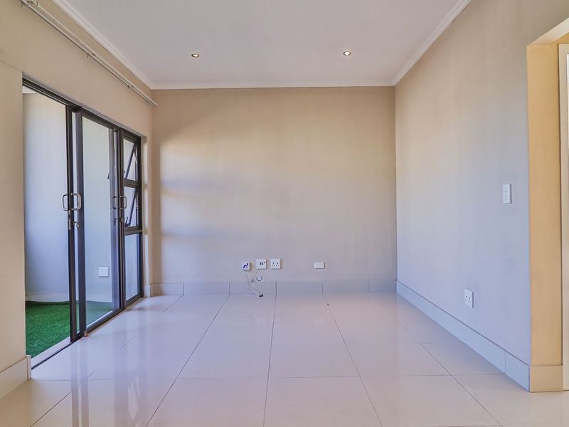 A bright, empty living room featuring neutral tiled flooring, recessed downlighting, and a sliding glass door providing access to an outdoor area.