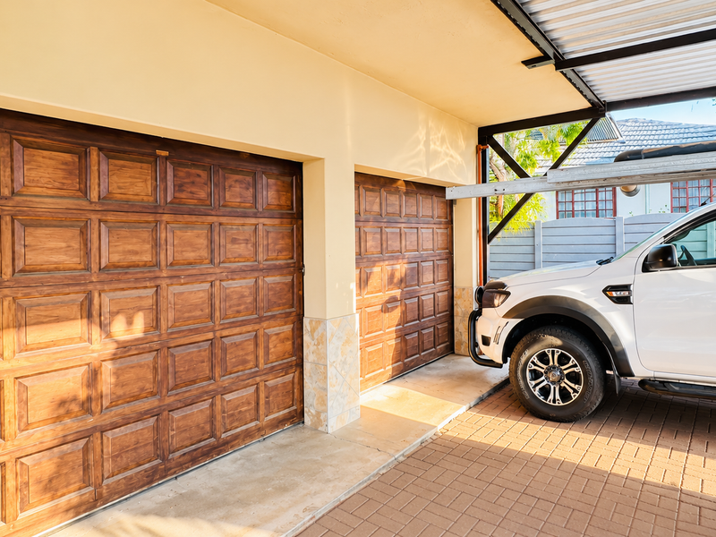 A well-maintained double garage featuring classic wooden doors and an adjacent covered carport area with paved flooring.