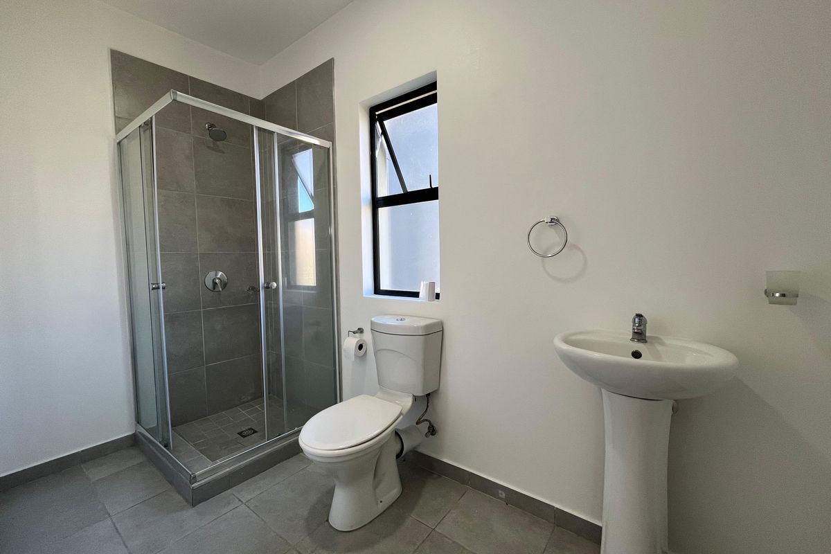 A clean, modern bathroom featuring a glass-enclosed shower, pedestal basin, and toilet with neutral grey floor tiles and white wall finishes.