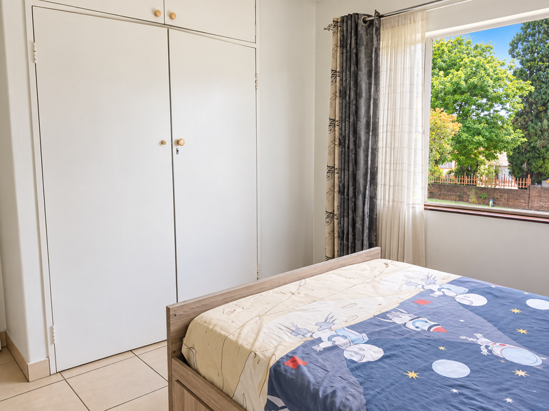 A bedroom featuring tiled flooring and a built-in wardrobe with overhead storage, offering natural light and a view of the garden.