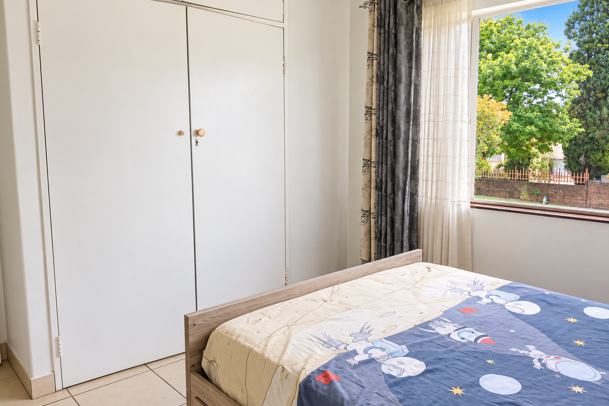 A bedroom featuring tiled flooring and a built-in wardrobe with overhead storage, offering natural light and a view of the garden.