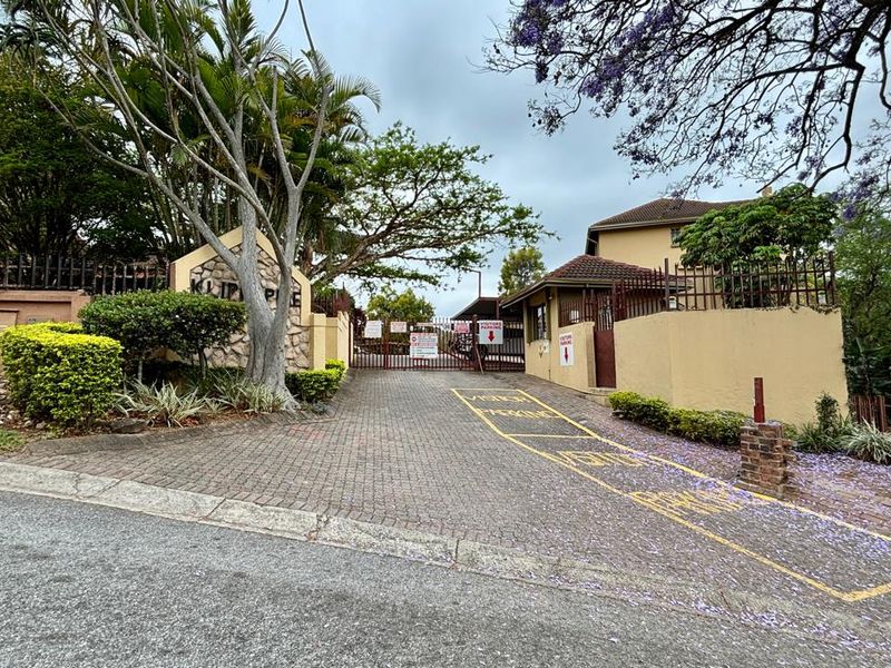The entrance to the apartment complex features a paved driveway, secure gated access, and perimeter walling with established landscaping.