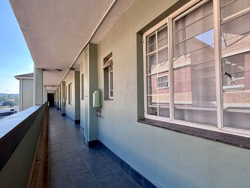 A covered exterior walkway or balcony featuring tiled flooring, security bars on windows, and a protective balustrade overlooking the building exterior.