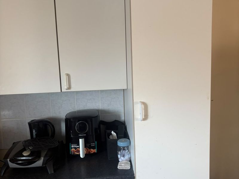 A compact kitchen area featuring white built-in cabinetry, dark laminate countertops, and a tiled backsplash, showing signs of wear and tear.