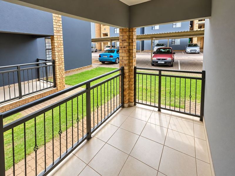 A clean, covered ground-floor patio featuring tiled flooring, brick support pillars, and a metal balustrade overlooking the complex parking area.