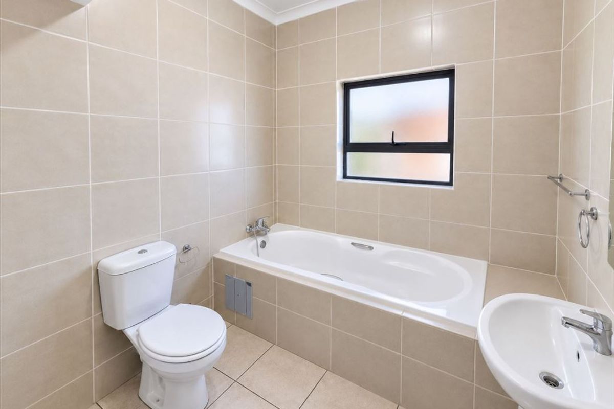 A clean, neutral-toned bathroom featuring a built-in bathtub, pedestal basin, and toilet with full-height wall tiling and a frosted window for privacy.