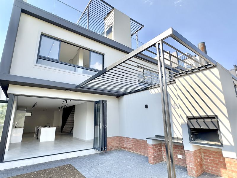 Rear view of a modern townhouse featuring a paved patio, built-in braai, metal pergola, and seamless indoor-outdoor flow through large bi-fold doors.
