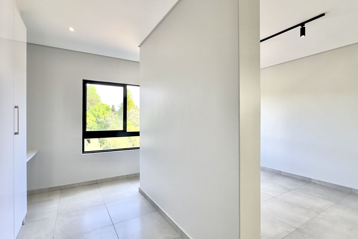 A bright, modern, and empty bedroom featuring neutral tiled flooring, built-in cupboards, and contemporary track lighting with large windows overlooking greenery.