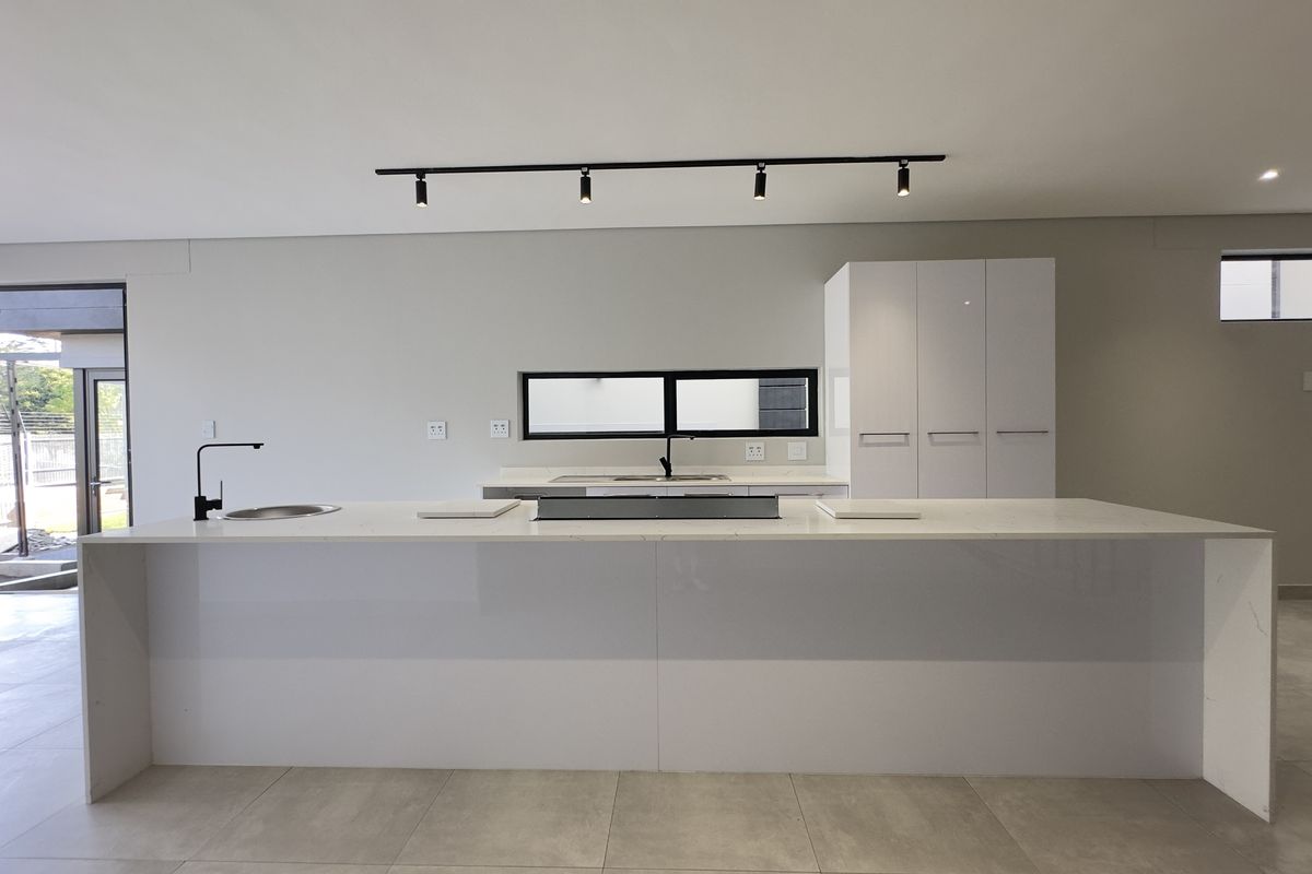 Modern, minimalist kitchen featuring a large central island with stone countertops, sleek white cabinetry, and contemporary track lighting in an open-plan layout.